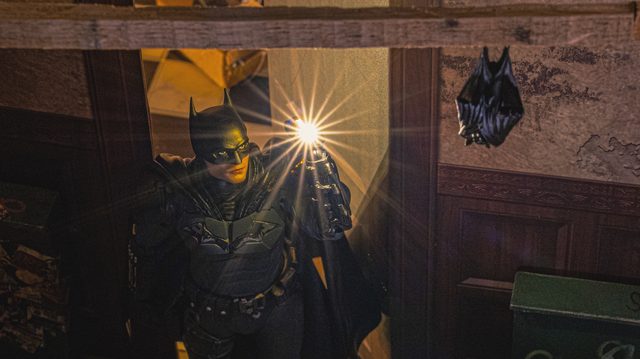 A Batman figure in a dark wooden room, a bright light flaring from his raised hand