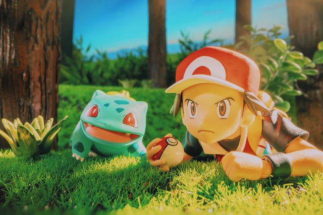 A Pokémon trainer figure lying in the grass, face to face with a Bulbasaur, a Poké Ball in hand