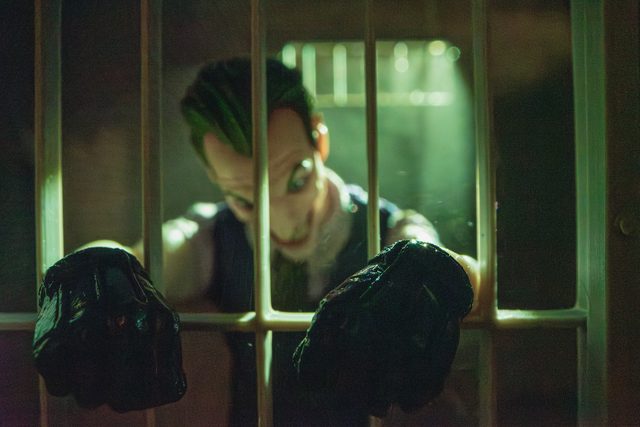 A green-haired figure gripping the bars of a cell, lit in green from behind
