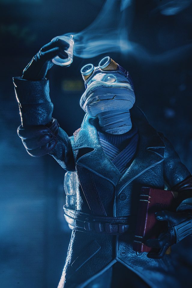 A bandaged figure in a trench coat and goggles holding up a smoking vial, in blue light