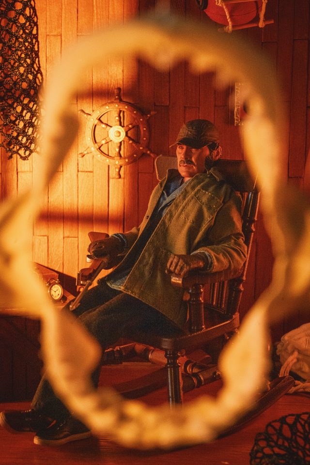 A fisherman figure in a rocking chair, seen through a shark’s jaws in a warm orange cabin